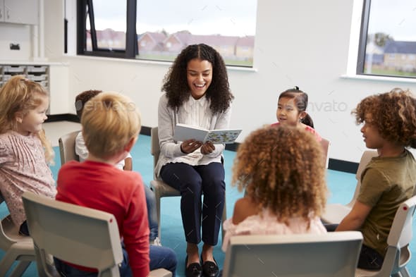 Young female school teacher reading a book to kindergarten children, sitting on chairs in a circle in the classroom listening